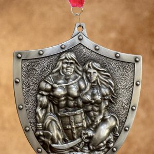 platinum pewter colored shield-shaped medal hanging from a red, white, and blue striped ribbon, featuring an embossed relief of a muscular warrior couple in heroic poses